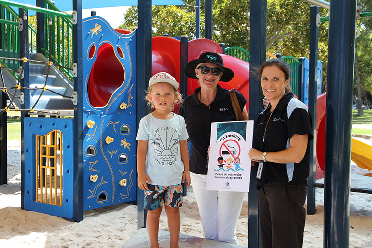 A young boy stands with two women in front of colourful playground equipment. One woman holds a certificate that reads Please do not smoke near our playgrounds.